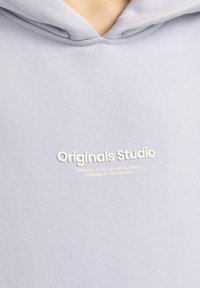 Svijetlo siva dukserica s kenguruh džepom, kapuljačom s vezicom i bijelim tekstom logotipa: "Originals Studio" na prednjoj strani. Meka tkanina.