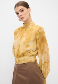 Fließende, gelbe Bluse mit hohem Kragen und langen Ärmeln. Verfügt über eine subtile marmorierte Textur und wird mit einem braunen Rock kombiniert.