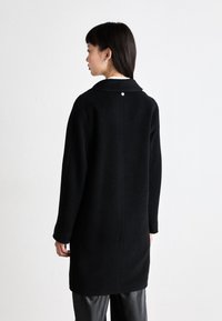 Schwarzer Mantel mit kragenförmigem Design, aus einem strukturierten Stoff gefertigt. Verfügt über einen einzelnen silbernen Knopf auf der Rückseite und lange Ärmel.