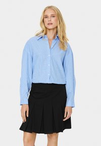 Camicia abbottonata a righe azzurro chiaro con sottili aperture, abbinata a una gonna plissè nera. La camicia presenta un colletto classico e maniche lunghe.