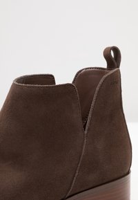 Detailní záběr na hnědou semišovou kotníkovou botu s koženým interiérem, zadním poutkem a dřevěným klínovým podpatkem na jednoduchém pozadí.