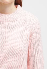 Pull en tricot rose clair avec un col haut et côtelé. Présente une maille lâche et des manches raglan, créant un aspect doux et texturé.