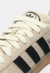 Tan suede sneaker with three dark blue velvet stripes, textured laces, and a round logo on the tongue. Rubber sole with light brown accents.