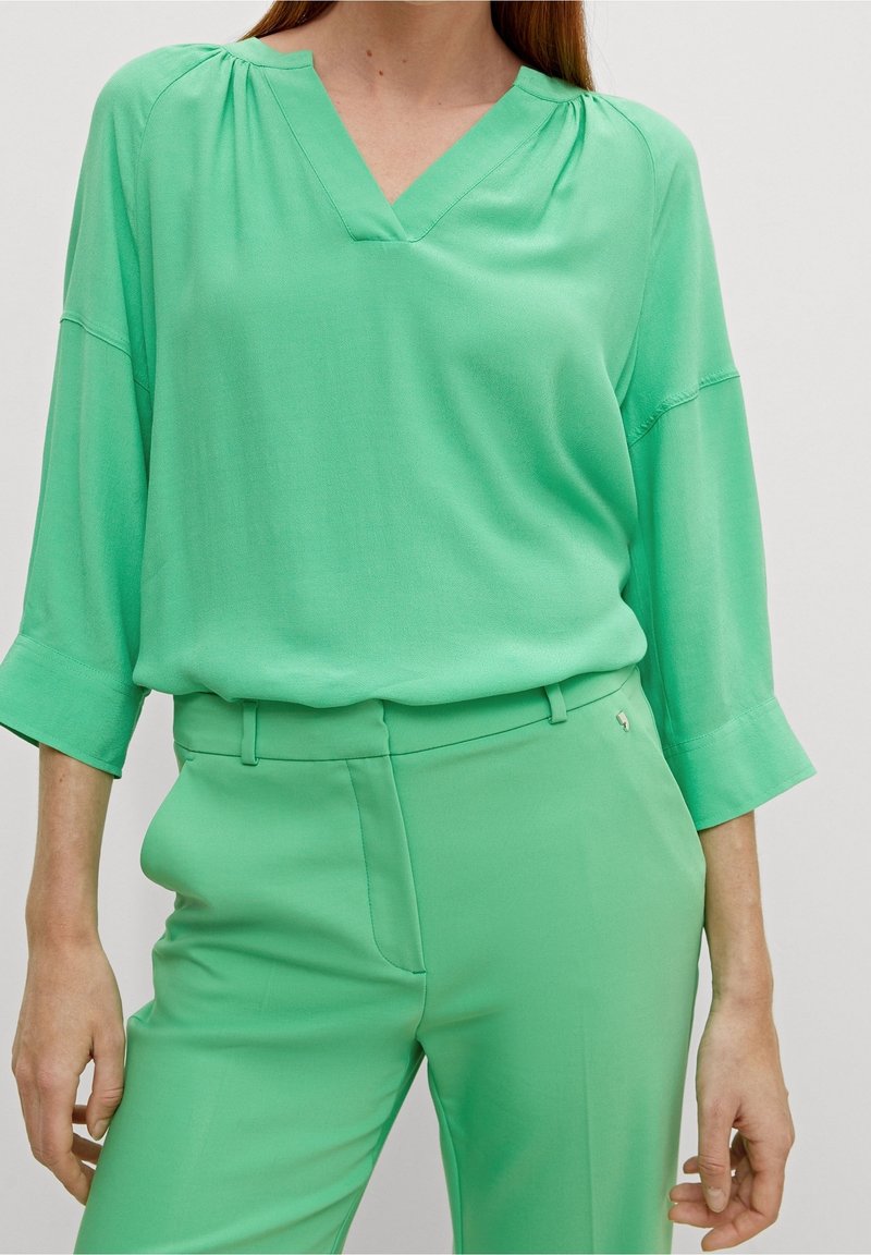 Femme portant un chemisier ample vert clair à col en V avec des manches 3/4 et un pantalon taille haute assorti contre un fond uni.