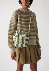 Streifenförmige grüne und weiße Stofftasche mit zwei Griffen und einem abnehmbaren Riemen, versehen mit einem Charm aus bunten Perlen und einem roten Druckdesign.