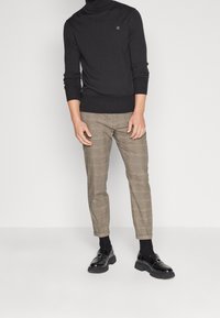 Black turtleneck sweater paired with plaid brown trousers. Trousers feature subtle tan accents. Footwear is black loafers with chunky soles.