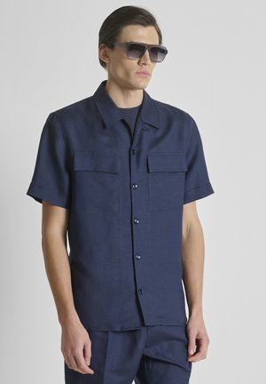 Homme portant une chemise bleu foncé à manches courtes boutonnée et un pantalon assorti, accessoirisé avec de grandes lunettes de soleil carrées noires, fond neutre.