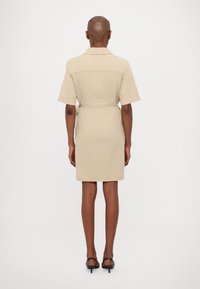 Personne portant une robe beige mi-longue à manches courtes avec une ceinture à la taille, debout, de dos, sur un fond clair uni.