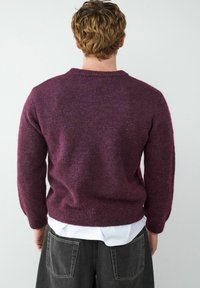 Lila Pullover aus einem strukturierten, weichen Strick. Mit einem runden Halsauschnitt und langen Ärmeln, kombiniert mit grauen weit geschnittenen Hosen.