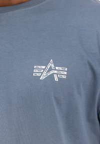 Camiseta de algodón azul de manga corta con un diseño gráfico texturizado en blanco que presenta líneas angulares y un símbolo abstracto en el pecho.