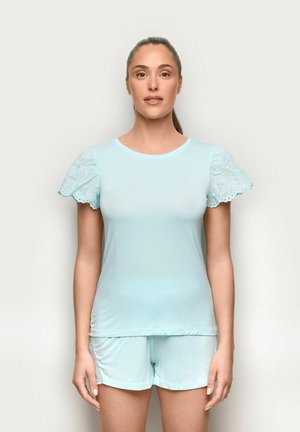Kobieta z włosami związanymi do tyłu, ubrana w jasnoniebieską bluzkę z krótkim rękawem z koronkowymi detalami i pasujące szorty, stojąca na jednolitym tle.
