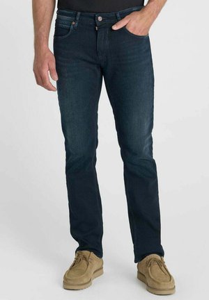 Mann trägt dunkelblaue Slim-Fit-Jeans und beige Wildleder-Freizeitschuhe, steht mit einer Hand in der Tasche vor einfachem Hintergrund.