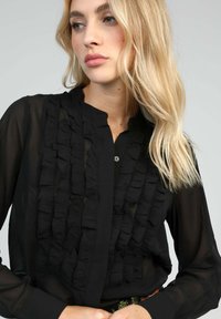 Blouse noire en tissu transparent, avec un détail froncé à l'avant, un plastron à boutons et des manches longues avec des poignets ajustés.