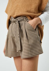 Pantalones cortos marrón de espiga con una cintura alta, pliegues doblados y un cinturón atado. Los bolsillos son visibles, combinados con un suéter de punto beige.