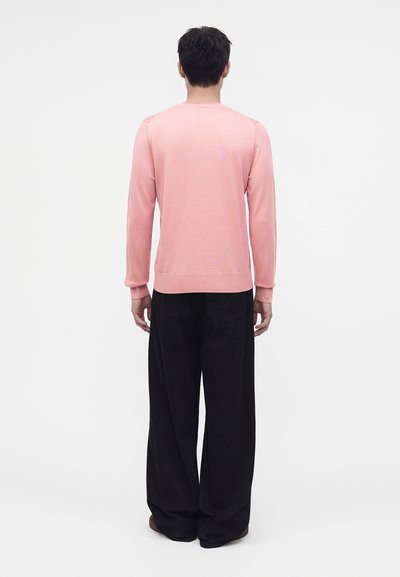 Homme debout de dos, portant un pull rose clair à manches longues et un pantalon noir ample sur un fond blanc uni.