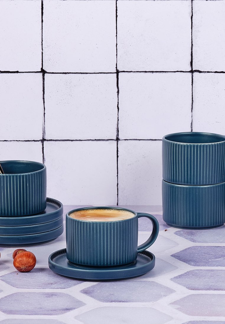 Tazza da caffè di ceramica blu scuro con texture rigata, piattino abbinato e due tazze impilate su una superficie decorata con nocciole vicine.