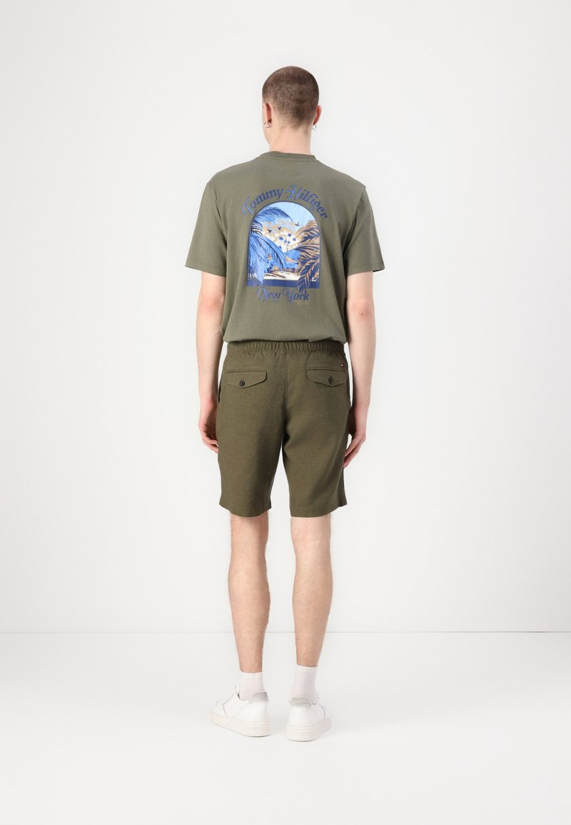 Tricou din bumbac verde cu un print grafic albastru pe spate, asortat cu șorturi verde oliv. Modelul poartă adidași și șosete albe.