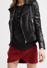 Giacca da motociclista nera in pelle con cerniere argentate e colletto a punta, abbinata a un top nero e a una gonna avvolgente rossa. Texture liscia.