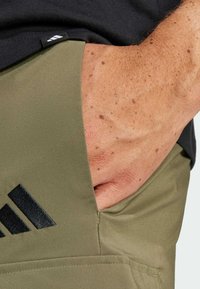 Olive green sports shorts featuring a black Adidas logo and a hand in the pocket, showcasing a smooth fabric texture.