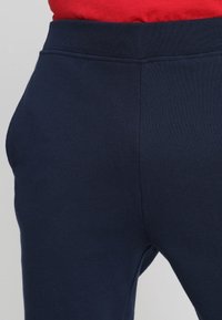Marineblauwe rekbroeken met een zachte textuur, voorzien van een tailleband en naaddetailing. Close-upweergave die de pasvorm en het materiaal benadrukt.