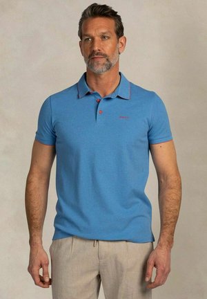 Man met kort donker haar en baard, die een lichtblauw poloshirt met korte mouwen en beige broek draagt tegen een neutrale achtergrond.