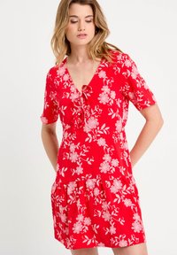 Robe florale rouge avec décolleté en V, manches courtes et ourlet rigolo. Fabriquée en tissu léger avec des motifs floraux blancs et un détail à nouer.