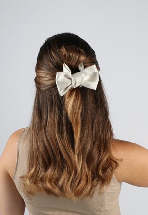 Satin-Haarbinde in Elfenbein, groß und strukturiert, getragen in einer halbhohen Frisur mit welligem braunem Haar, die das glänzende Material und die feinen Falten präsentiert.