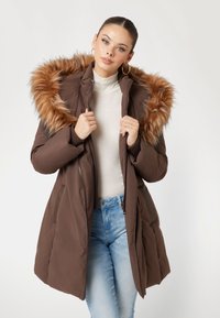 Rudas šiltas paltas su dirbtinio kailio gobtuvu, užtrauktuku, šoninėmis kišenėmis ir vėduoklės formos rankovėmis, dėvimas ant šviesaus golfuko ir mėlynų džinsų.