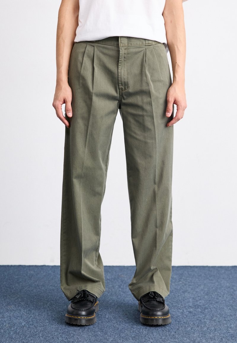 Pantalons plissés vert olive en coton, présentant une coupe décontractée et une taille mi-haute. Associés à des chaussures noires, sur un tapis bleu.