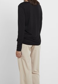 Černý top s dlouhými rukávy a kulatým výstřihem, s nařaseným designem vzadu. V kombinaci se širokými béžovými kalhotami, s hladkou texturou po celé ploše.