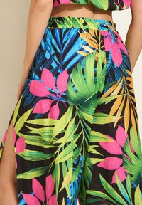 Spodnie mają czarną tkaninę z intensywnymi, tropikalnymi wzorami liści w kolorze zielonym, różowym i żółtym, elastyczny pas oraz boczne rozcięcia.