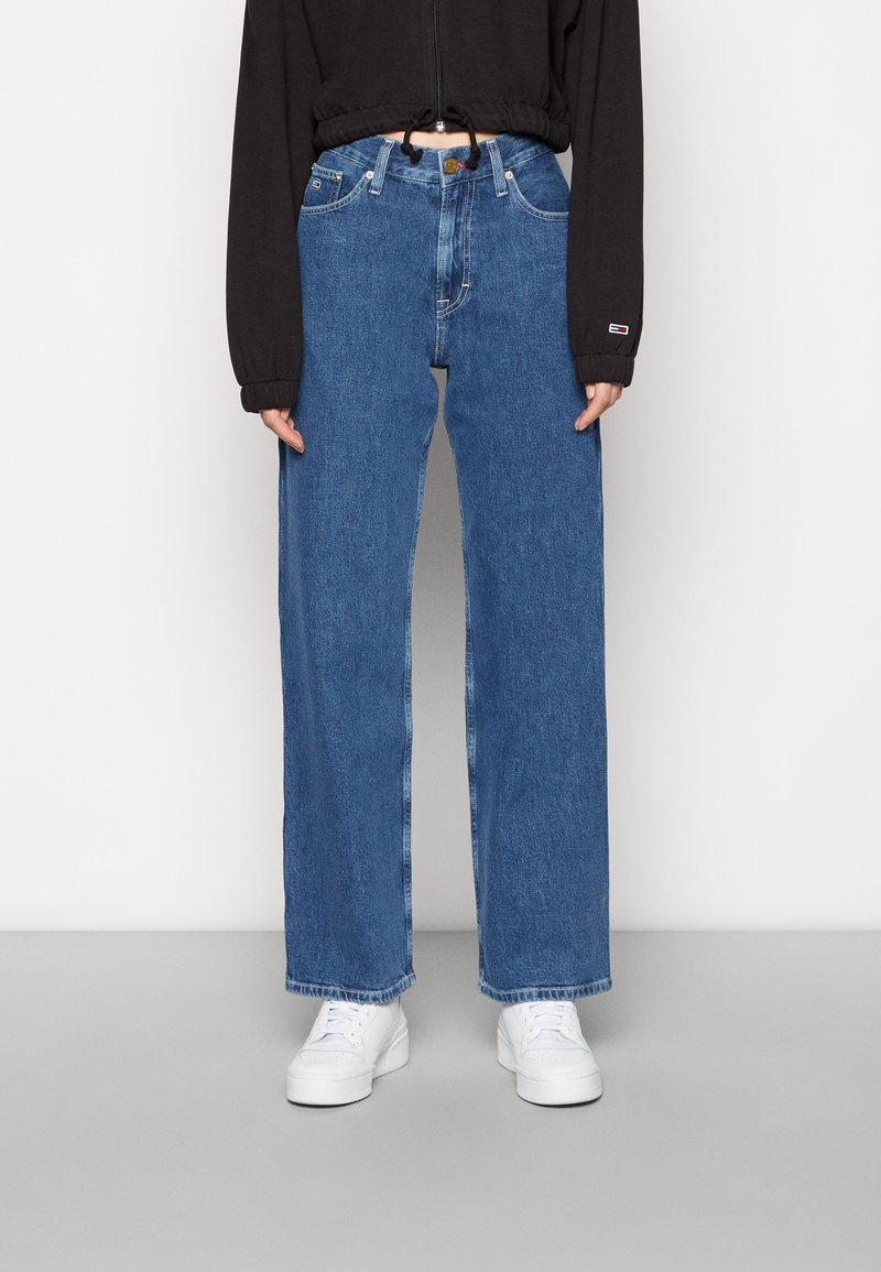 Jeans de pierna ancha en azul claro con cintura alta, que cuentan con cinco bolsillos y un cierre de botón, combinados con zapatillas blancas y una chaqueta corta oscura.