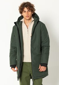 Groene gewatteerde jas met een capuchon, voorzien van een ritssluiting aan de voorkant, zijzakken en elastische manchetten. Draagt over een lichtbeige trui.