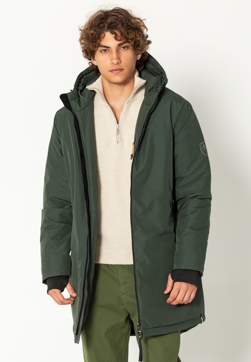 Groene gewatteerde jas met een capuchon, voorzien van een ritssluiting aan de voorkant, zijzakken en elastische manchetten. Draagt over een lichtbeige trui.