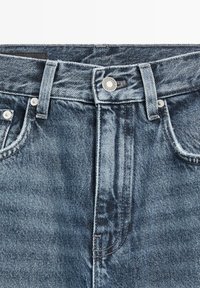 Close-up van de tailleband van een blauwe spijkerbroek met knoop, riemlussen, voorzakken en zichtbare stikdetails.