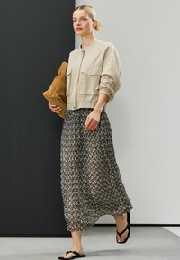 Beige lyhyempijakkunen, jossa on kaksi taskua, yhdistetty musta-valkoiseen kuosilliseen maxi-hameeseen ja mustiin flip-floppeihin, pitäen ruskeaa clutch-laukkua.