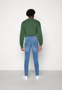 Zelený pletený svetr, modré úzké džíny s zadními kapsami a bílé tenisky. Oblečení má casual design s moderním vzhledem.