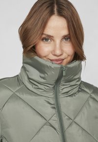 Hellgrüne, gesteppte Jacke mit hohem Kragen und Frontreißverschluss. Der Stoff hat eine glatte, glänzende Textur und ein rautenförmiges Muster.