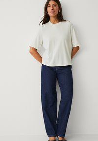 Haut blanc à manches courtes avec un col rond, associé à un jean large bleu foncé. Le modèle se tient avec les mains dans les poches, sur un fond neutre.