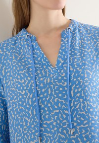 Gros plan sur le cou et le haut de la poitrine portant un haut à col en V bleu à motif avec des cordons réglables et des embouts métalliques.