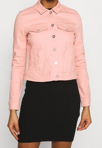 Chaqueta de mezclilla en rosa claro con cierres de botones, dos bolsillos en el pecho y un diseño ligeramente corto. Llevada sobre una falda negra acanalada.