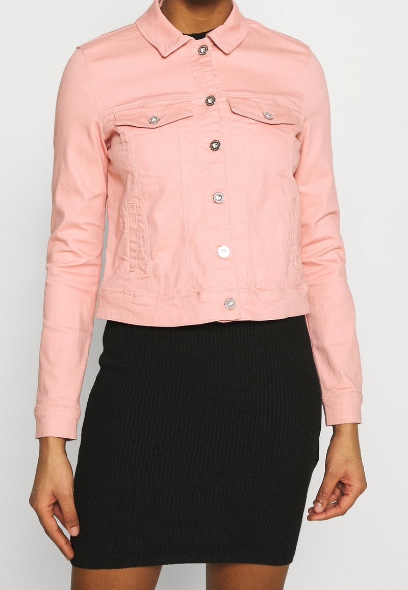 Chaqueta de mezclilla en rosa claro con cierres de botones, dos bolsillos en el pecho y un diseño ligeramente corto. Llevada sobre una falda negra acanalada.