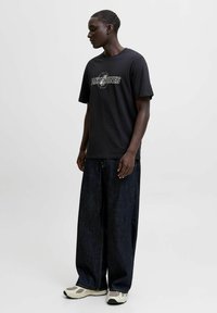 Noor mees seisab vasakule vaatades, seljas must "Jack & Jones" t-särk, laiad tumehalli teksad ja valged tossud ühetaolisel taustal.