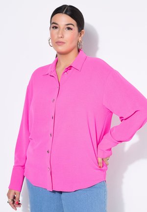 Frau mit dunklem Haar, trägt Creolen, rosa Bluse mit Knöpfen, hellblaue Jeans, steht mit einer Hand in der Hüfte vor weißem Hintergrund.
