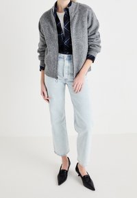 Szara dzianinowa kurtka bomberka z zapięciem na zamek, noszona na koszuli w kratę zapinanej na guziki, w połączeniu z jasnoniebieskimi prostymi dżinsami i czarnymi szpilkami.