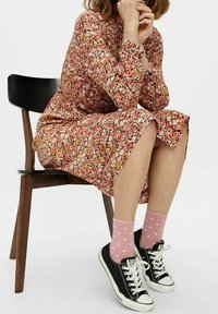 Abito floreale in arancione, rosa e bianco che arriva sopra il ginocchio. Scarpe Converse nere con suola bianca e calzini a pois rosa. Sfondo con una sedia di legno.