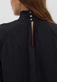 Black blouse with a high neck, featuring a keyhole opening and three buttons at the back. Fabric appears soft and textured.