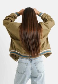 Kaki bomberjack met gestreepte ribgebreide manchetten en zoom, gecombineerd met lichtblauwe high-waisted jeans. Lange steile haren zichtbaar.