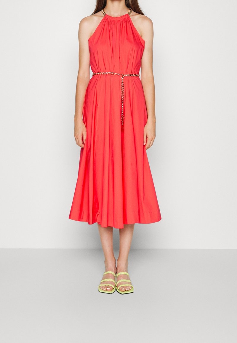 Robe corail à encolure halter avec un tissu froncé et une ceinture en chaîne métallique. Silhouette évasée atteignant le mollet. Portée avec des sandales à lanières jaunes.