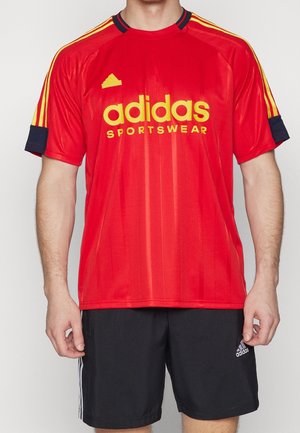 Man draagt een rood Adidas Sportswear-shirt met gele strepen en zwarte Adidas-short met witte strepen aan de zijkanten.